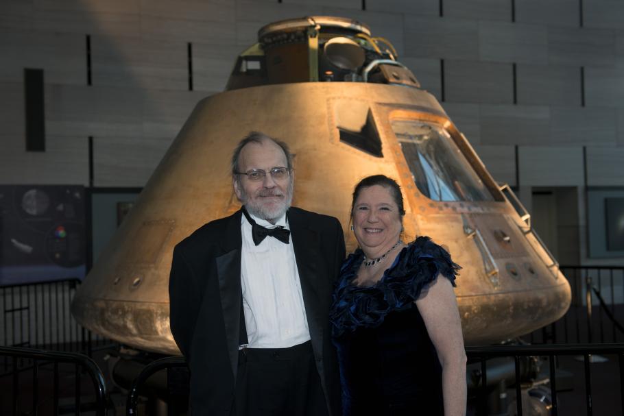 Two people, a white male and female, pose in front of the Apollo Command Module, a conic-shaped metal spacecraft.